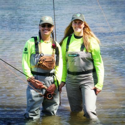 Two outdoor guides for Virginia State Parks fly fishing at Shenandoah River State Park. The guides are standing in the river and holding fly fishing poles and wearing waders and fishing gear.