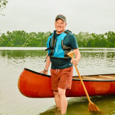 Senator Tim Kaine standing in river holding canoe with paddle and wearing personal flotation device. Paddling or canoeing and camping on James River in Virginia.