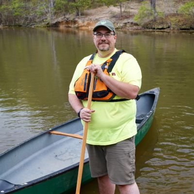 Sammy Zambon, Visitor Experience Specialist for Virginia State Parks standing in water at Lake Anna State Park next to a canoe, holding a paddle, and wearing a life jacket pfd.