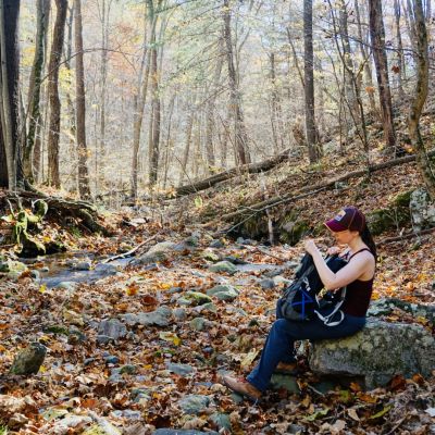 Woman sits on a rock with hiking pack near a stream in the Blue Ridge Mountains during autumn.