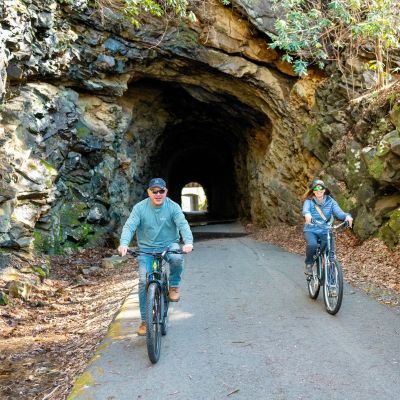 Big Stone Gap Town Manager, Steve Lawson, and podcast host, Jessica Bowser, ride bikes side-by-side through a stone tunnel on the Powell River Trail, a popular rail-to-trail.