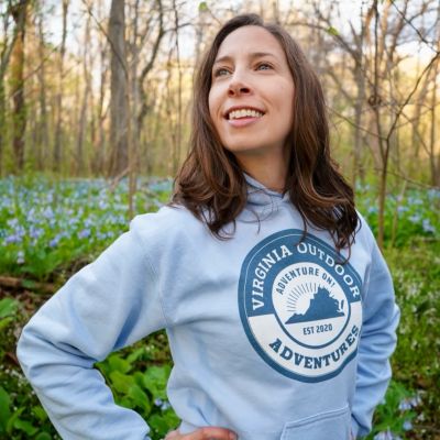 Podcast host, Jessica Bowser, stands among Virginia Bluebell wildflowers in spring, wearing blue hoodie with Virginia Outdoor Adventures logo.